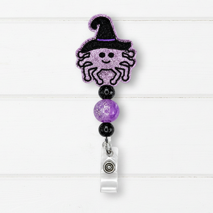 A Halloween-themed retractable badge reel featuring a glittery purple spider wearing a black witch hat, separated by purple and black beads from a clear plastic clip on a white wood plank background.