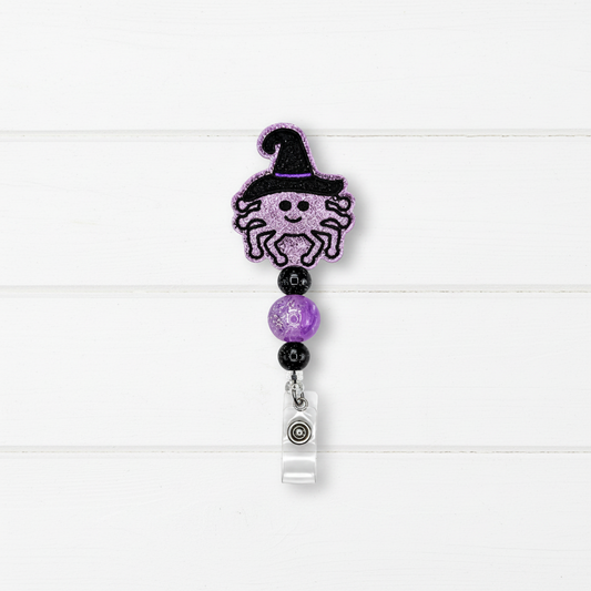 A Halloween-themed retractable badge reel featuring a glittery purple spider wearing a black witch hat, separated by purple and black beads from a clear plastic clip on a white wood plank background.