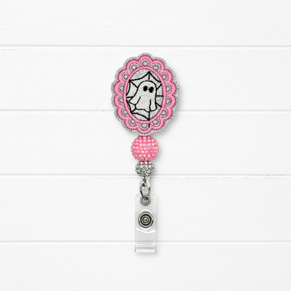 A pink and silver embroidered badge reel with a ghost design, shown on a white wood plank background.