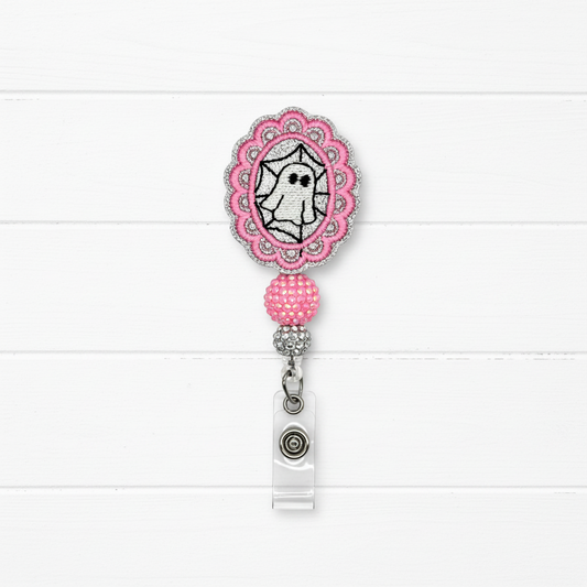 A pink and silver embroidered badge reel with a ghost design, shown on a white wood plank background.