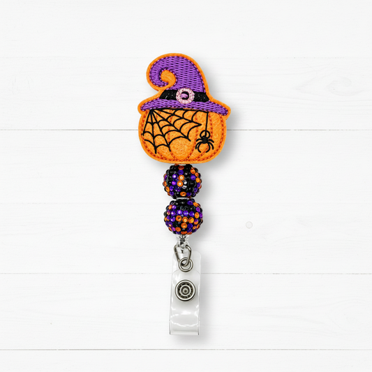 A Halloween-themed retractable badge reel featuring an embroidered orange pumpkin with a purple witch hat, black spiderweb pattern, and a small black spider on the front. The reel is adorned with two purple and orange beaded spheres and has a clear plastic clip at the bottom.