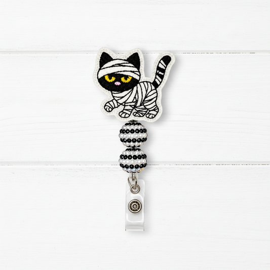 A Halloween-themed retractable badge reel featuring a mummy cat design. The cat is embroidered in black and white with bright yellow eyes, wrapped in white bandages.