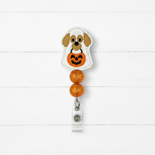 A Halloween-themed embroidered badge reel featuring a white ghost with tan ears and a black face holding an orange pumpkin, attached to two orange glitter beads and a silver clip on a white wood plank background.
