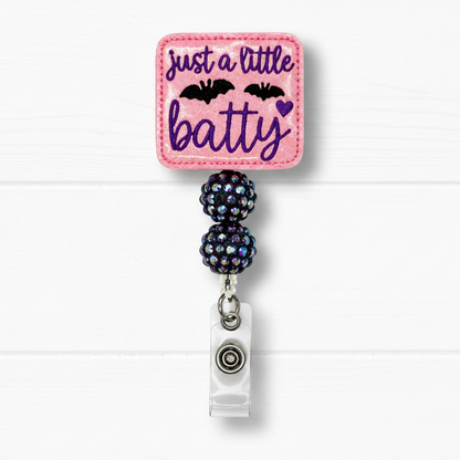A pink embroidered badge reel with the text 'just a little batty' in purple script, decorated with two black bats and a small purple heart. The pink square patch is attached to two dark iridescent rhinestone-covered beads and a silver metal clip, set against a white wood plank background.