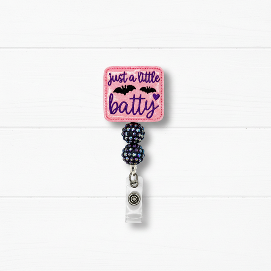 A pink embroidered badge reel with the text 'just a little batty' in purple script, decorated with two black bats and a small purple heart. The pink square patch is attached to two dark iridescent rhinestone-covered beads and a silver metal clip, set against a white wood plank background.