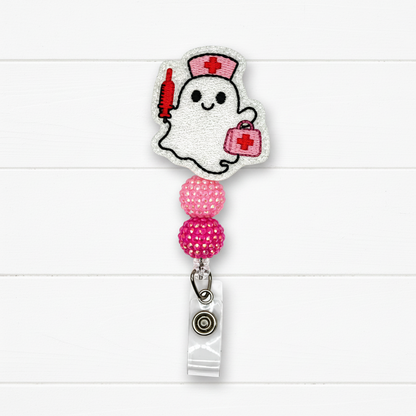 A ghost-shaped embroidered badge reel cover featuring a white ghost with a red nurse hat, red syringe, and pink first-aid kit, attached to a retractable clip with pink rhinestone beads on a white wood background.