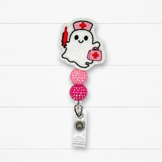 A ghost-shaped embroidered badge reel cover featuring a white ghost with a red nurse hat, red syringe, and pink first-aid kit, attached to a retractable clip with pink rhinestone beads on a white wood background.