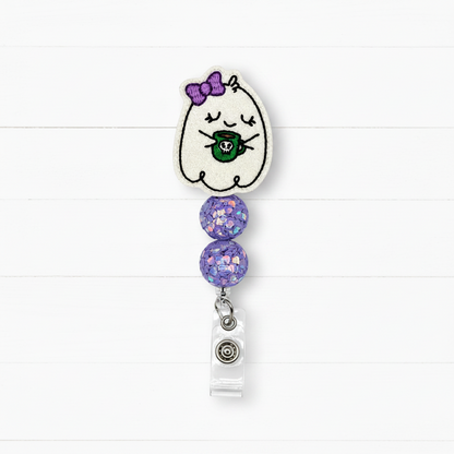 A ghost-themed retractable badge reel featuring a white embroidered ghost with a purple bow and green mug, accented with two purple glitter beads and a clear plastic clip.