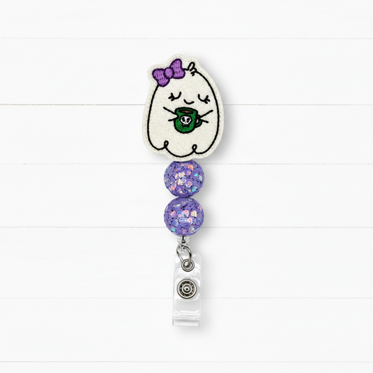 A ghost-themed retractable badge reel featuring a white embroidered ghost with a purple bow and green mug, accented with two purple glitter beads and a clear plastic clip.