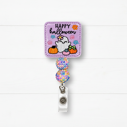 A Halloween-themed retractable badge reel with a lavender embroidered patch featuring a ghost, pumpkins, flowers, and the text 'HAPPY halloween.' The patch is attached to two glittery multicolor beads and a clear plastic clip with a silver metal snap, shown against a white wood plank background.