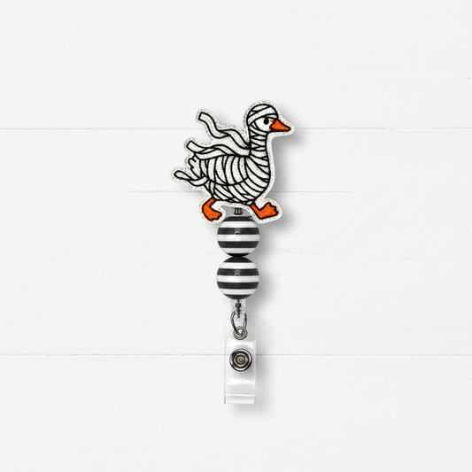 A novelty retractable badge reel featuring a whimsical cartoon duck with black-and-white striped body bands and bright orange feet and beak, mounted above a black-and-white striped roller ball and white clip on a white wood plank background.