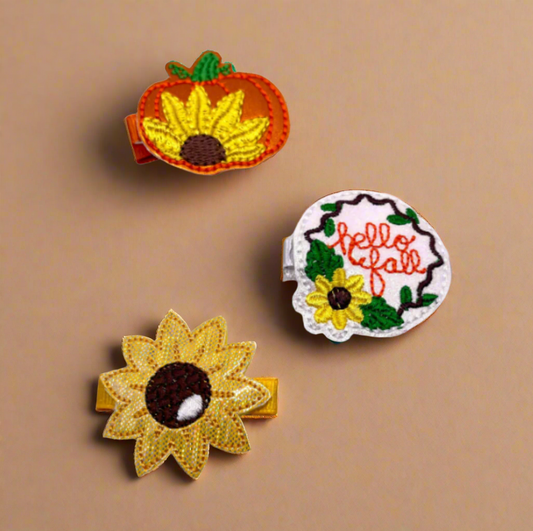 A set of three hair clips with fall-themed designs including a sunflower, a pumpkin with a sunflower, and the text 'Hello Fall' on a green and white patterned background.