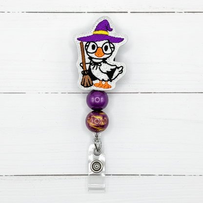 A Halloween-themed retractable badge reel featuring a white penguin dressed as a witch with a purple hat and broom, two purple beads, and a silver metal clip on a white wood plank background.
