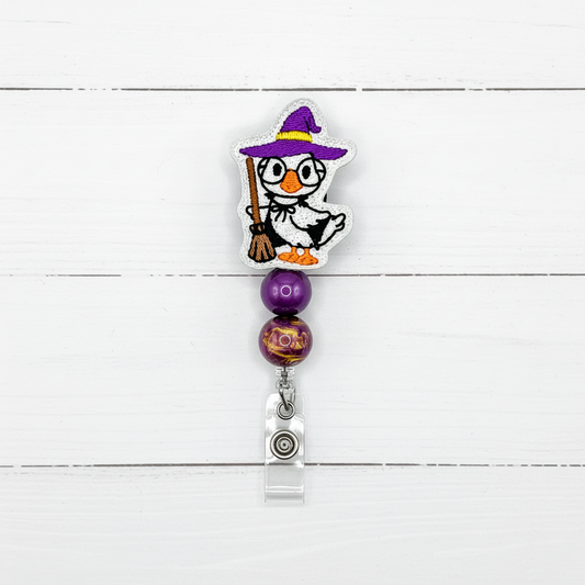 A Halloween-themed retractable badge reel featuring a white penguin dressed as a witch with a purple hat and broom, two purple beads, and a silver metal clip on a white wood plank background.