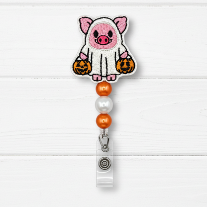 A Halloween-themed embroidered badge reel clip featuring a cute pink pig dressed in a white ghost with orange jack-o'-lanterns, hanging from orange and pearl beads on a white wood plank background.