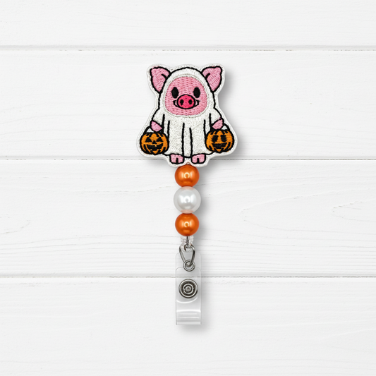 A Halloween-themed embroidered badge reel clip featuring a cute pink pig dressed in a white ghost with orange jack-o'-lanterns, hanging from orange and pearl beads on a white wood plank background.