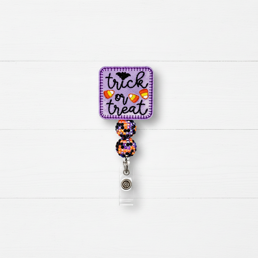 A Halloween-themed retractable badge reel with a purple square patch that reads 'trick or treat' in black script, decorated with a bat, candy corn, and orange-and-white candy corn motifs. The attached retractable strap has two glittery purple, orange, and black pom-poms and a silver metal clip on a white wood plank background.