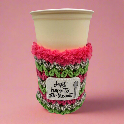 A crocheted multicolored cup cozy with text 'Just here to stir the pot' displayed on a beige cup.