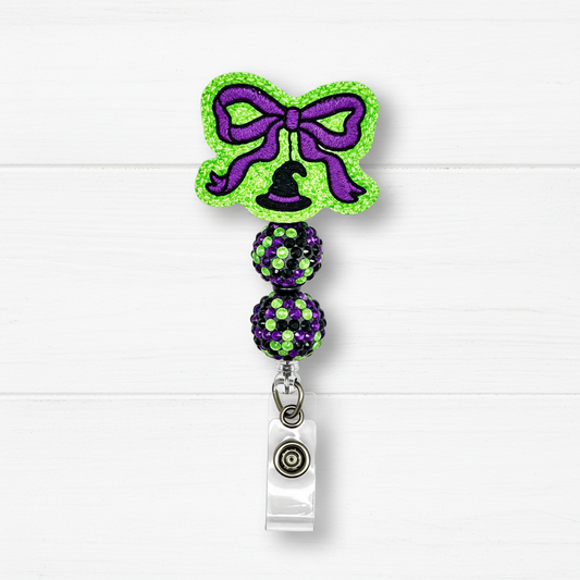 Halloween-themed retractable badge reel with a green glitter bow, purple glitter accents, a black witch hat, and two glittery purple and green beads on a clear plastic clip, shown on a white wood background.