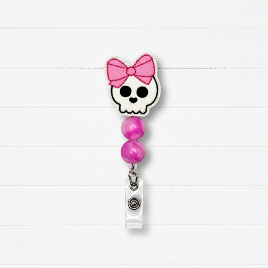 A retractable badge reel with a skull and pink bow design, featuring two pink beads and a silver clip, set against a white wood plank background.