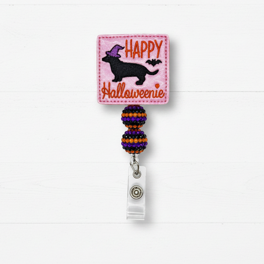 A Halloween-themed retractable badge reel featuring a pink square patch with a black dachshund wearing a purple witch hat, orange 'HAPPY' text, and 'Halloweenie' script. The reel has purple, orange, and black beaded loops and a silver metal clip, shown against a white wood plank background.