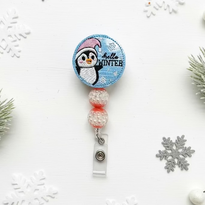Retractable badge holder with penguin design on a white background with snowflakes and pine branches.