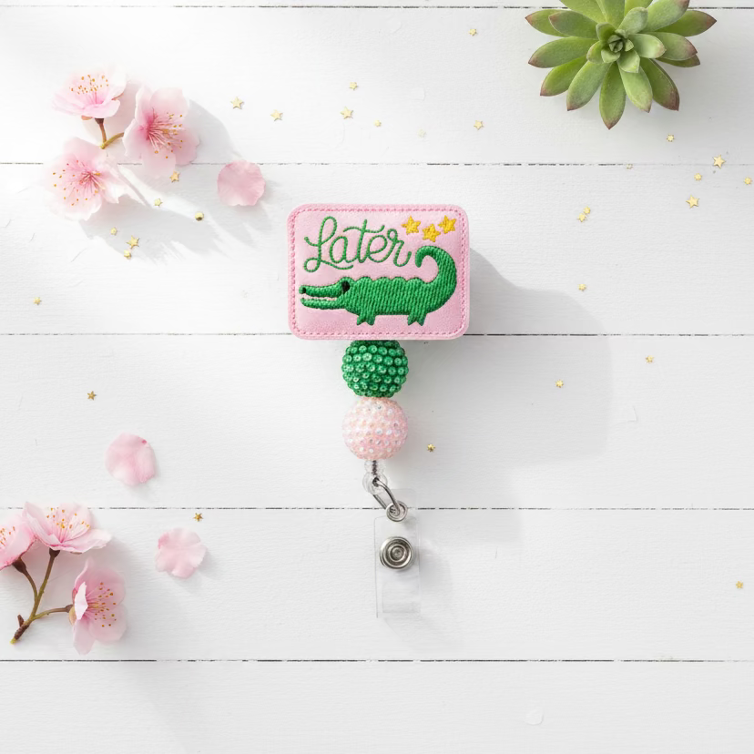 A pink embroidered patch with a green alligator and the word 'Later' in script, attached to a retractable badge reel with green and iridescent beads on a light pink background.