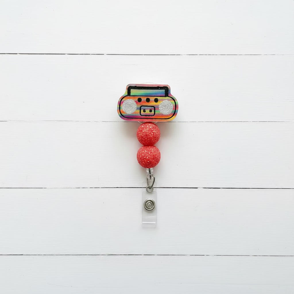 A novelty retractable badge reel featuring a colorful, glittery boombox design with two red glitter beads and a silver clip, shown on a light pink background.