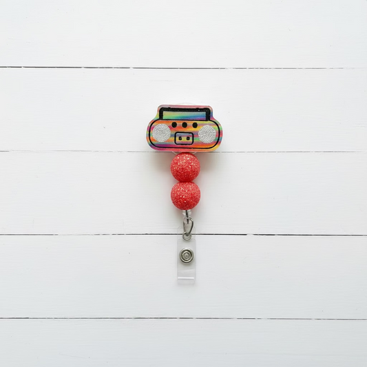 A novelty retractable badge reel featuring a colorful, glittery boombox design with two red glitter beads and a silver clip, shown on a light pink background.