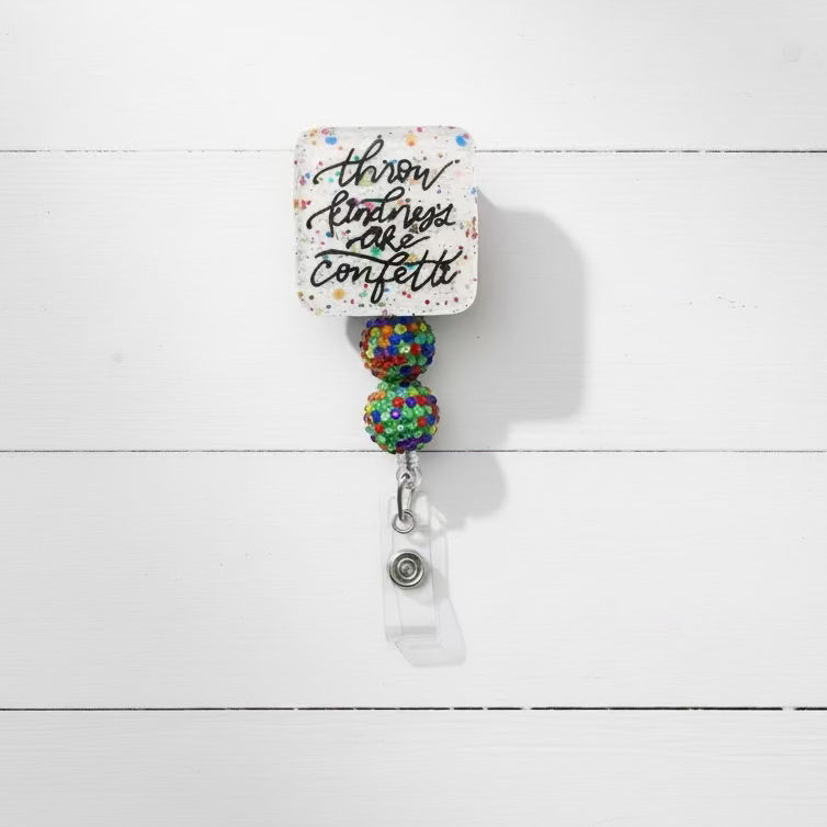 A square white badge reel with a glittery multicolor confetti background and the phrase 'throw parties are confetti' in black script. The reel is attached to two round, glittery multicolor beads and a silver metal clip on a light lavender background.