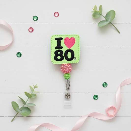 A square embroidered badge reel featuring a bright lime green background with 'I' and '80' in black, a pink heart, and a beaded pink and green loop with a silver clip.