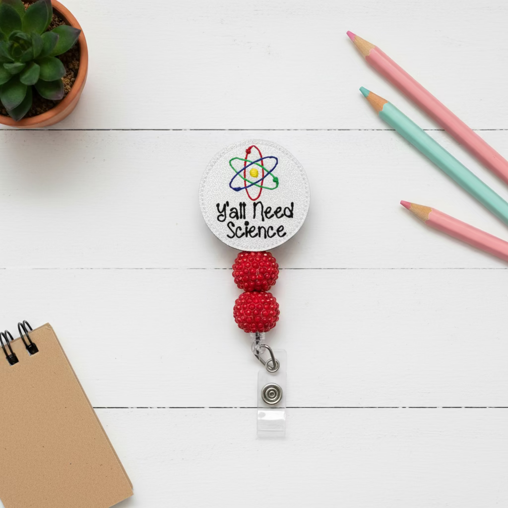 A circular white retractable badge reel featuring the text 'Y'all Need Science' and a colorful atom graphic, with two red beaded loops and a clear plastic clip below, shown on a light pink background.