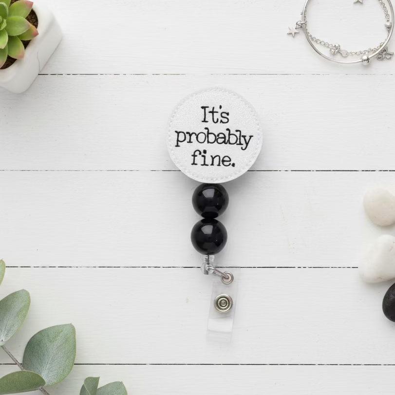 A novelty retractable badge reel with a round off-white fabric top that reads 'It's probably fine.' in black text, attached to two black beads and a silver clasp with a clear plastic backing.