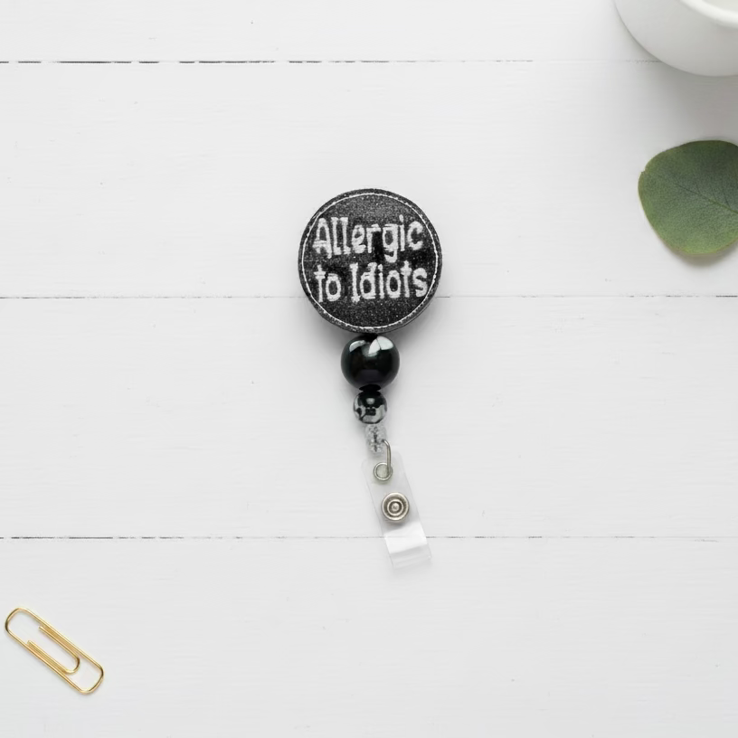 A circular black patch with white embroidered text reading 'Allergic to Idiots' hangs from a black and white marbled bead, a silver lobster clasp, and a clear plastic badge reel on a light pink background.