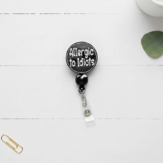 A circular black patch with white embroidered text reading 'Allergic to Idiots' hangs from a black and white marbled bead, a silver lobster clasp, and a clear plastic badge reel on a light pink background.