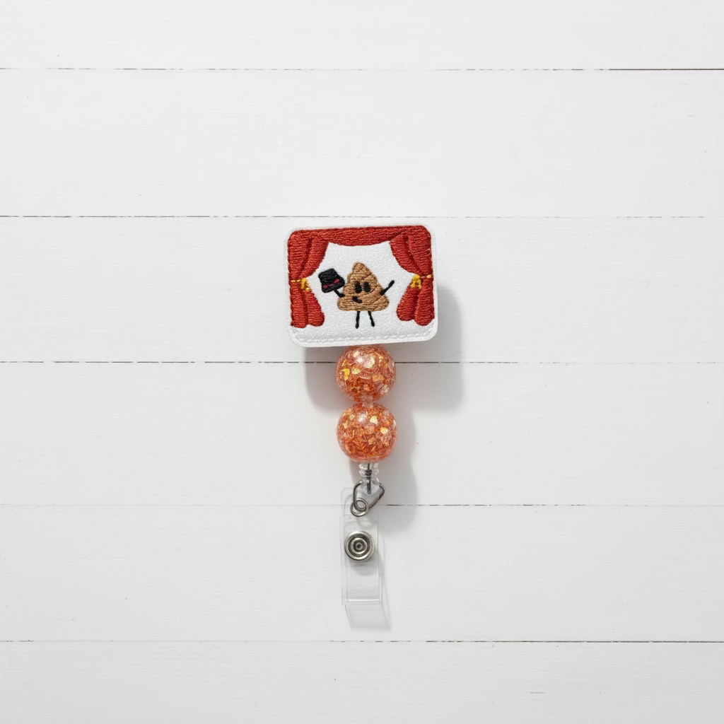 A novelty retractable badge reel featuring a white embroidered top with red curtains and a brown poop emoji wearing a small black top hat. Two round orange glitter beads hang below, connected to a silver metal clip holding a clear plastic sleeve with a metal reel insert.
