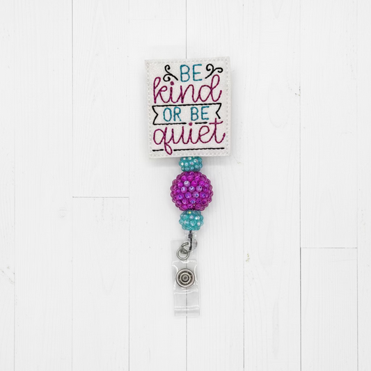 A fabric swatch card with the embroidered text 'BE kind OR BE quiet' in teal and magenta, attached to a purple beaded ball and a silver charm on a light pink background.
