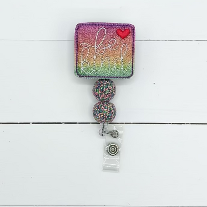 A square glitter badge reel cover with a rainbow gradient background, silver glitter script text, a small red heart, and two dangling glitter-covered beads attached to a clear plastic clip.