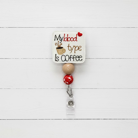 An embroidered badge reel featuring a square cream patch with the text 'My blood type is COFFEE' in black and red, alongside a small coffee cup graphic. The patch hangs from a tan and red bead chain with a silver metal clasp and clear plastic sleeve, photographed on a plain white background.