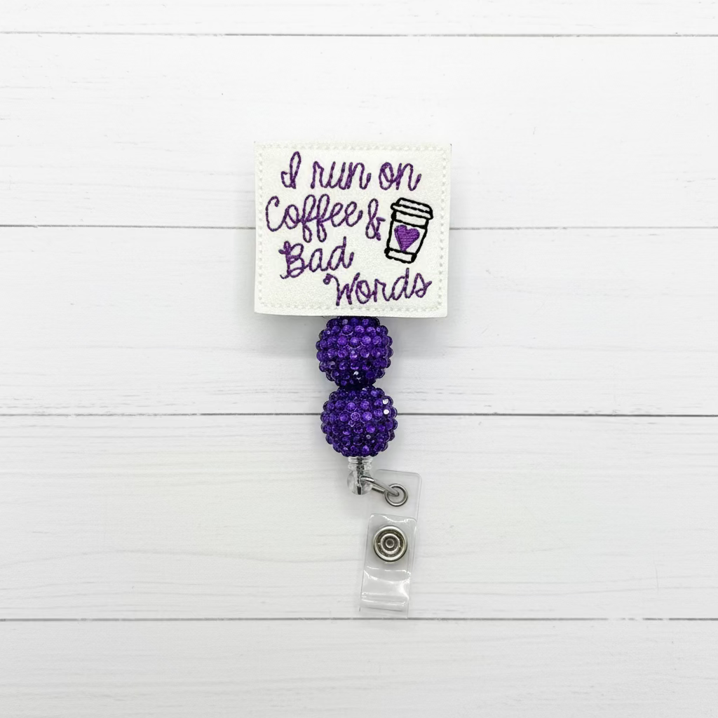 A white embroidered badge reel cover with the phrase 'I run on Coffee & Bad Words' in purple script and a small coffee cup graphic, attached to two purple rhinestone-covered balls and a clear plastic clip.