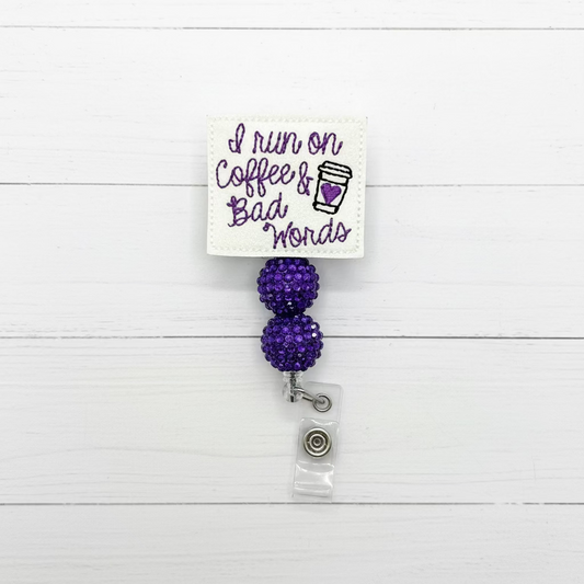 A white embroidered badge reel cover with the phrase 'I run on Coffee & Bad Words' in purple script and a small coffee cup graphic, attached to two purple rhinestone-covered balls and a clear plastic clip.