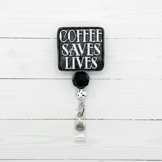 A square black sign-style badge reel with the phrase 'COFFEE SAVES LIVES' in distressed white lettering, attached to a faceted black bead, white pearl, and clear clip on a white background.