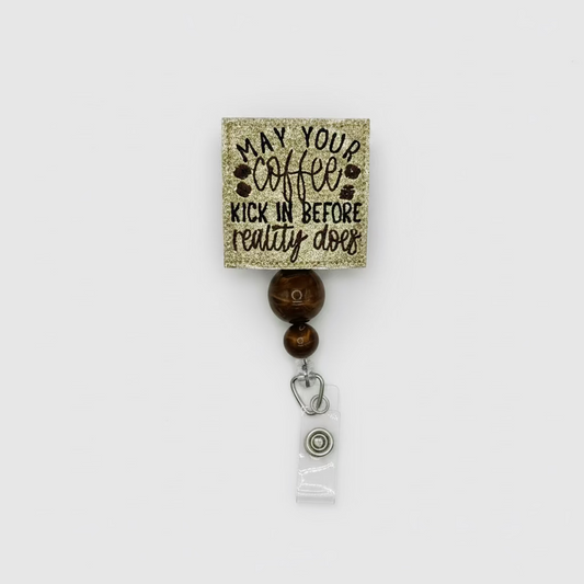 A square gold glitter badge reel with a printed coffee-themed quote reading 'MAY YOUR coffee KICK IN BEFORE reality does' in black lettering. The reel is attached to two brown tiger-eye style beads and a silver metal clip holding a clear plastic ID badge holder, shown on a plain white background.