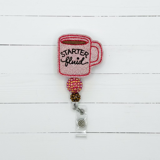 A novelty retractable badge reel featuring a glittery pink mug with 'STARTER fluid' written in black script, attached to a clear plastic ID clip with a metal snap button.