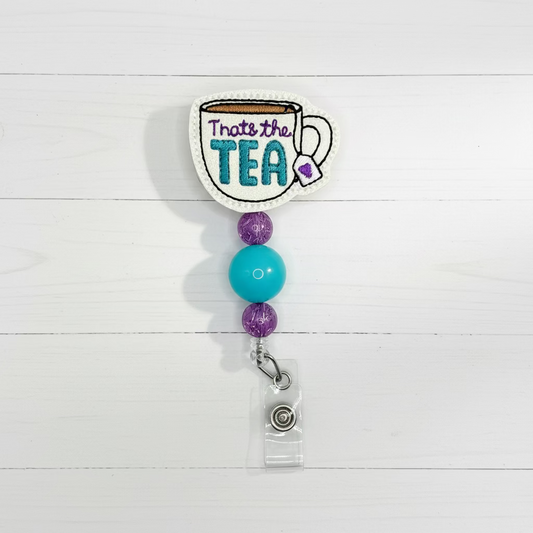 An embroidered patch shaped like a teacup with the words 'That's the TEA' in purple and teal, hanging from a clear plastic retractable badge reel with purple and turquoise beads on a white background.