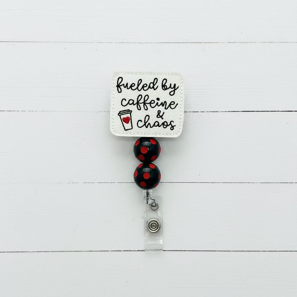 A square white embroidered badge reel with the phrase 'fueled by caffeine & chaos' in black script and a small red heart icon, attached to two black beads with red polka dots and a silver clasp on a white background.