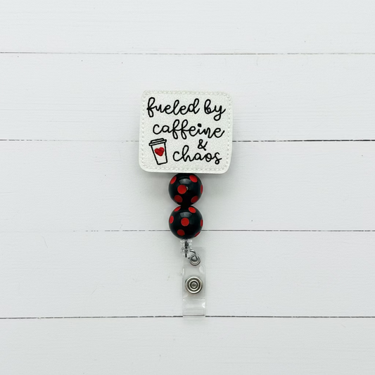 A square white embroidered badge reel with the phrase 'fueled by caffeine & chaos' in black script and a small red heart icon, attached to two black beads with red polka dots and a silver clasp on a white background.