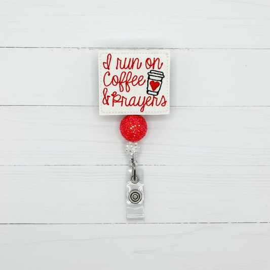 A square embroidered patch with the phrase 'I run on Coffee & Prayens' in red script and a coffee cup graphic, attached to a red glitter ball and retractable badge reel with a small charm, on a plain light background.