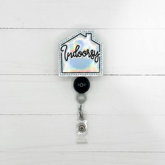 A house-shaped badge reel with an iridescent holographic background, black script text reading 'Indoorsy', a black round bead, a silver spacer, and a clear plastic clip on a white background.
