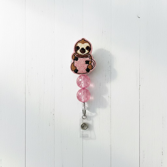 A pink retractable badge reel with a cute sloth holding a pink glittery mug, suspended by two pink faceted beads on a silver clip.